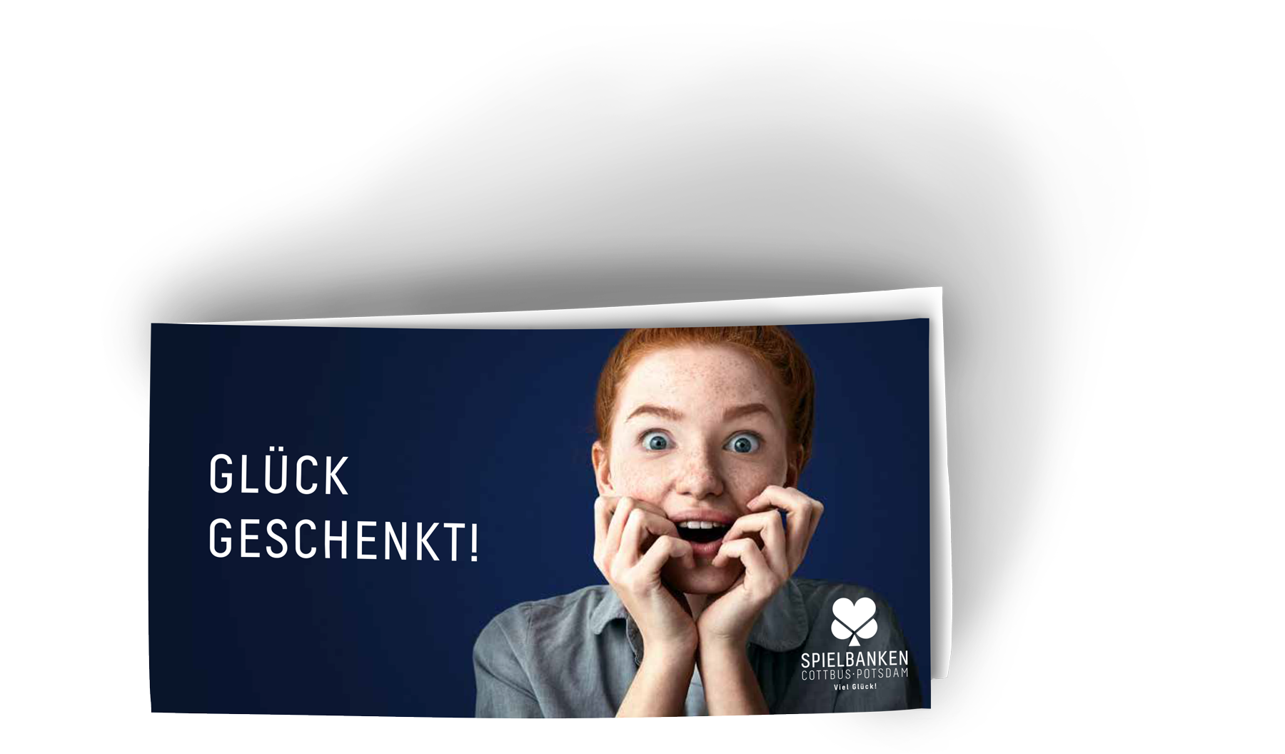 Zu sehen ist ein Foto des Gutscheins für die Spielbanken Potsdam und Cottbus. Das zusammengeklappte Format des Gutschein ist Din lang. gefalzt ist er an der linken kurzen Seite. Er ist blau und darauf abgebildet eine junge Frau mit roten, zu einem Dutt hochgebunden Haaren, die beide Hände freudig überrascht an den leicht geöffneten Mund legt. Auf dem Gutschein steht links in Großbuchstaben Glück Geschenkt.